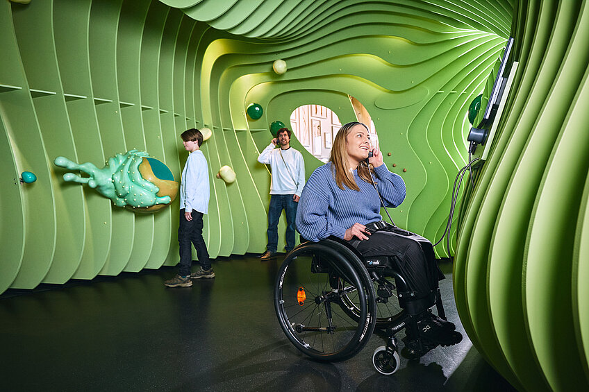 Ein Junge und zwei Erwachsene stehen in dem überdimensionalen Modell einer grünen Pflanzenzelle. Der junge berührt eine grüne Zellorganelle. Der stehende Mann und die im Rollstuhl sitzende Frau halten Einhandhörer an ihre Ohren und lauschen. 