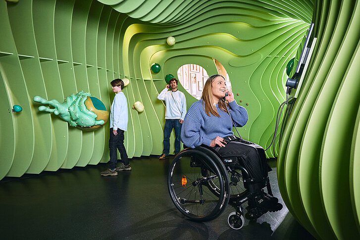 Ein Junge und zwei Erwachsene stehen in dem überdimensionalen Modell einer grünen Pflanzenzelle. Der junge berührt eine grüne Zellorganelle. Der stehende Mann und die im Rollstuhl sitzende Frau halten Einhandhörer an ihre Ohren und lauschen. 