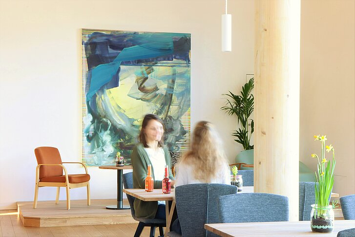 Im Bistro des NAWAREUMs sitzen zwei Frauen an einem Tisch und unterhalten sich. Die Möbel sind aus verschiedenfarbigem Holz und Polstern. Im Hintergrund hängt ein abstraktes Gemälde mit Blau- und Gelbtönen.