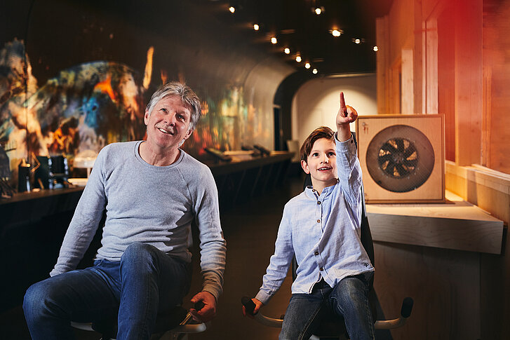Ein Junge und sein Opa sitzen auf Ergometern im NAWAREUM. Sie treten in die Pedale. Der Junge zeigt nach oben und sein Opa schaut hinterher. Im Hintergrund sieht man das Modell eines Windkanals.