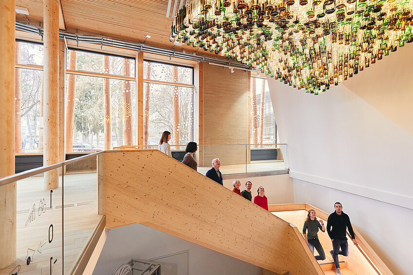 Im Eingangsbereich des NAWAREUMs gehen Gäste im Rahmen einer Führung die Treppe hinunter. Die Architektur besteht hauptsächlich aus Holz und großen Fenstern. An der Decke hängt ein Kunstwerk aus hunderten beleuchteten Glasflaschen.