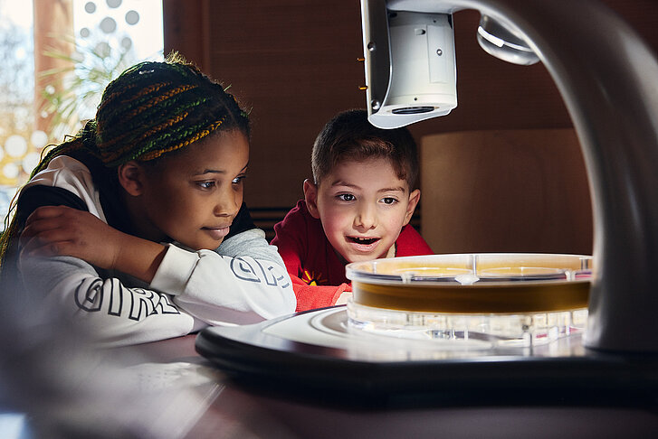 Zwei Kinder sitzen vor einem hell beleuchteten Mikroskop und schauen gespannt auf den Objektträger.