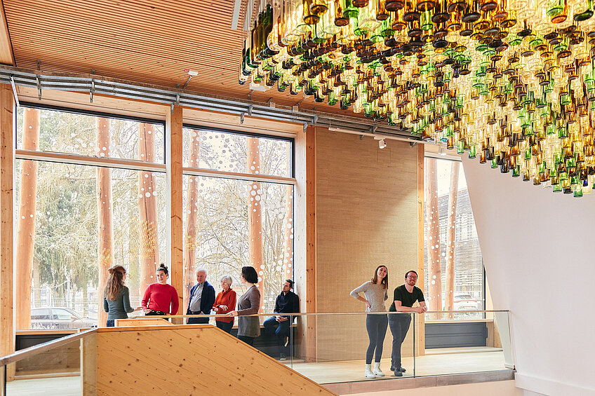 Im Eingangsbereich des NAWAREUMs lauschen Gäste einer Führung. Die Architektur besteht hauptsächlich aus Holz und großen Fenstern. An der Decke hängt ein Kunstwerk aus hunderten beleuchteten Glasflaschen.